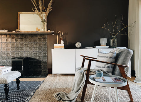 A living room featuring a wood and grey wool chair in the foreground and white sideboard in the background. The walls are painted dark brown.