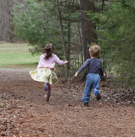 two kids running through nature- boy and girl, with sticks in the woods, dessed in a dress and trousers