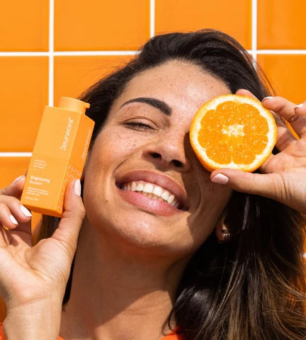 Woman with bright happy skin in orange bathroom, holding Vitamin C serum and orange fruit