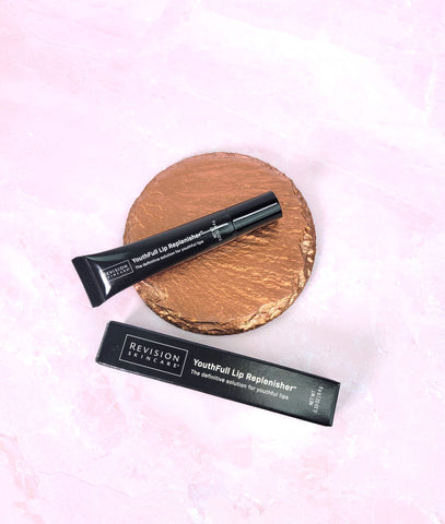 A tube of Revision Skincare Youthfull Lip Replenisher with its box, lying on a copper circle, above a pink marbled background
