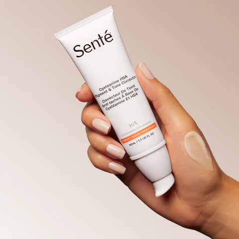 A person's hand, holding a tube of Sente Cysteamine HSA Pigment & Tone Corrector.