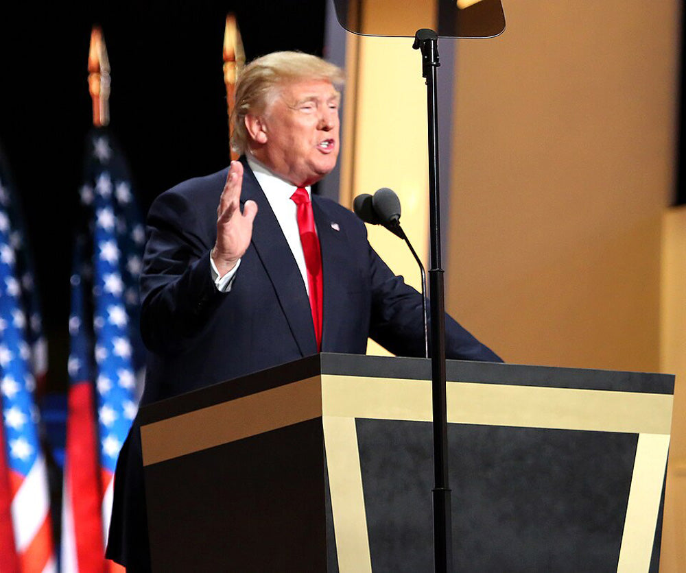 Donald Trump giving a speech at a podium with teleprompters, gesturing with his hands in front of American flags.
