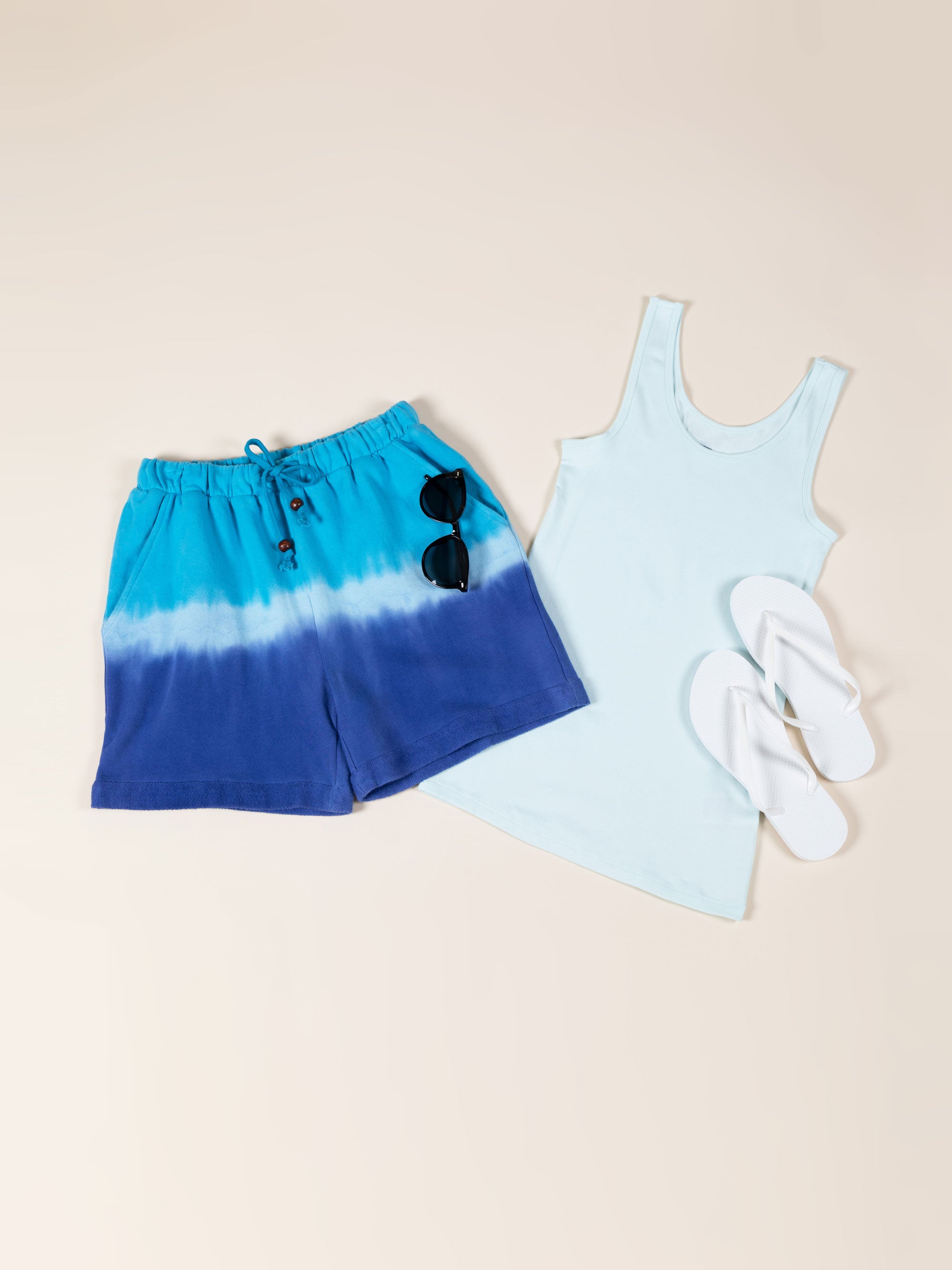 A pair of hand-dyed shorts are positioned to the left of a brightly-colored tank top and white flip-flops