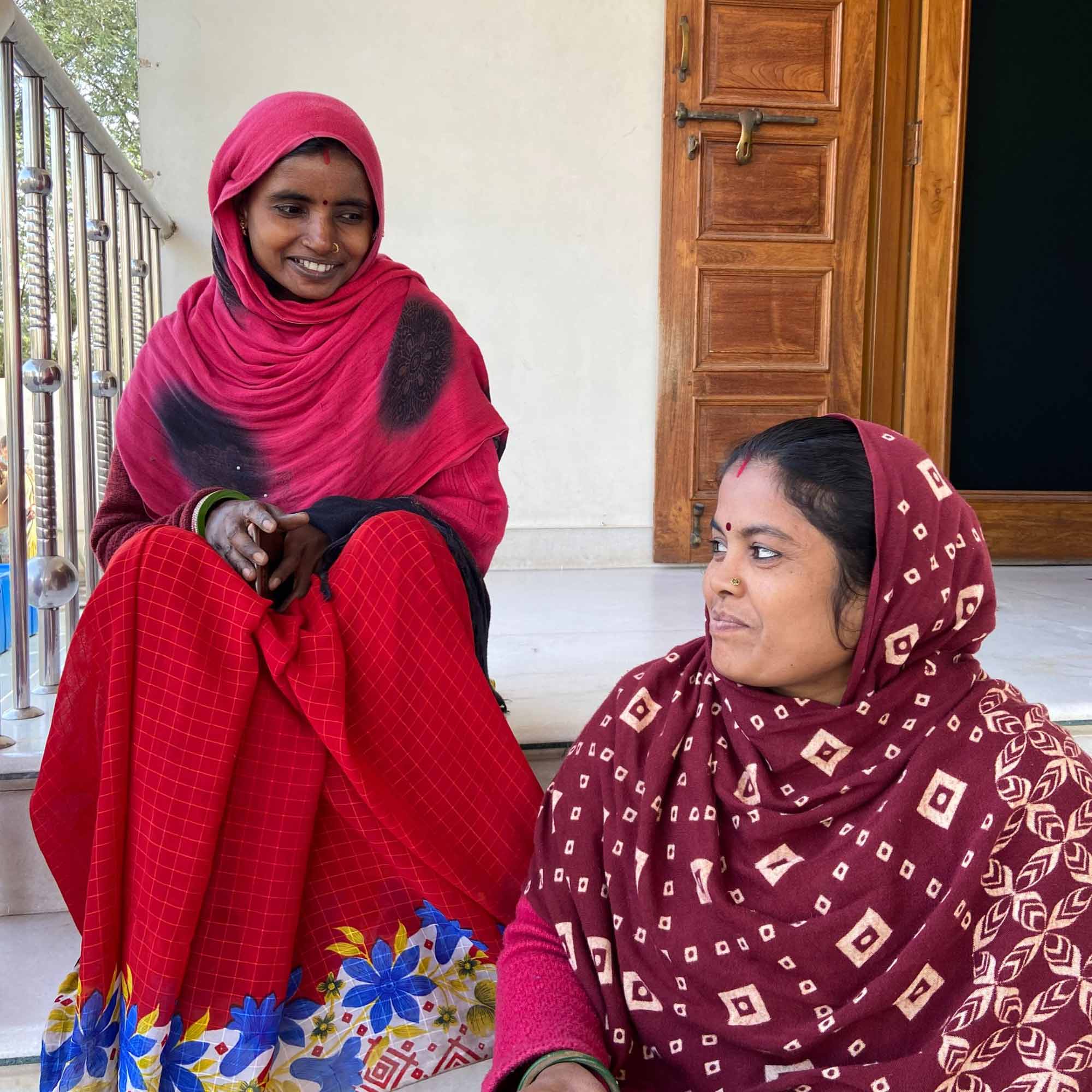 Two artisan women sit on the steps outside of a building and smile while they chat.d