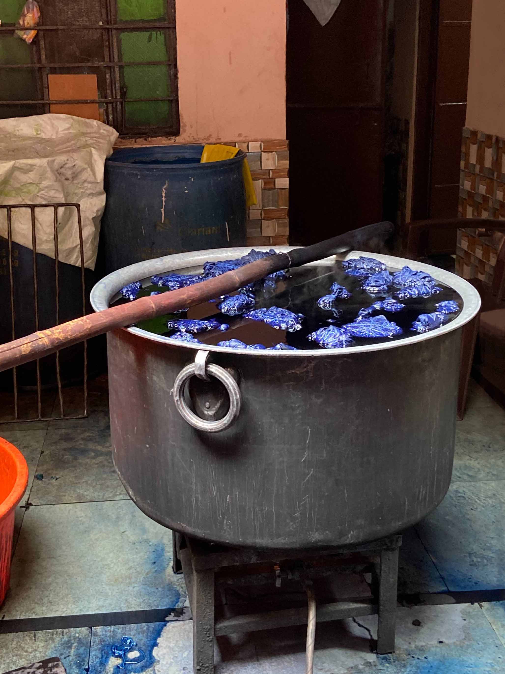 A very large, metal pot sits in the middle of a room. Water, dye, and farbic fill the pot which is stirred by a long wooden stick.