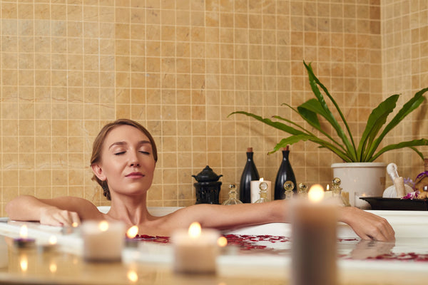 A woman sits in a bathtub filled with water and rose petals surrounded by burning candles