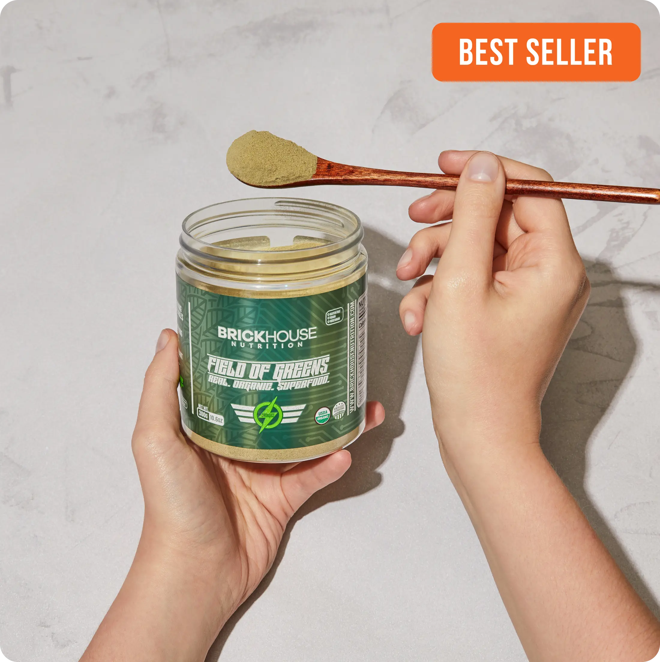 Hands holding an open jar of 'Field of Greens' superfood powder with a wooden spoon.