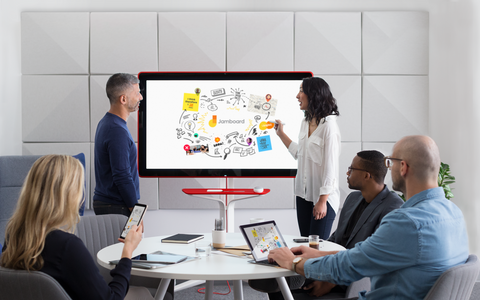 A photograph of a meeting space with two people standing in front of a Jamboard. They both have a stylus in their hand and are collaborating on a Jam. In front of them, there are two people looking at the same Jam from their laptop and mobile device.