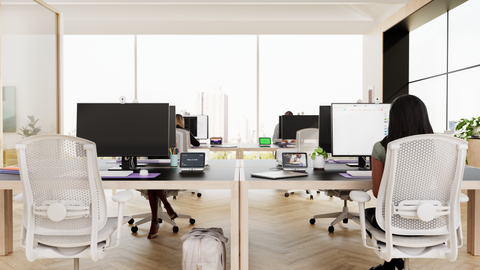 A photograph of an office space with multiple desks and chairs. Some desks are occupied whilst others are vacant. Each desk features a screen, webcam, keyboard, mouse, and a Logi Dock Flex.