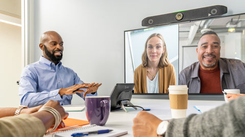 An image of a meeting space with people seated around a table. At the front of the room there is a TV screen displaying a video conferencing call in which two people are present remotely. Mounted on top of the screen is the Poly Studio X52 video bar. On the table, there is a Poly TC10 touch controller which is facing towards a man at the front of the room.