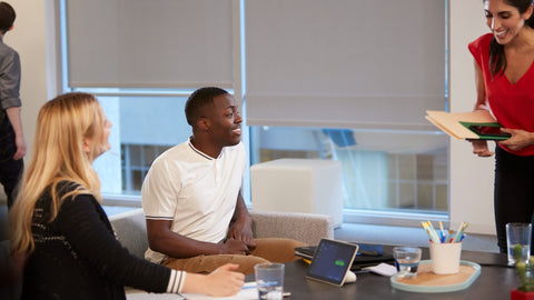 An image of a meeting space. Two people are sitting on chairs at the table whilst another person is standing up as they get ready to join a call. On the table, a Poly TC8 can be seen.