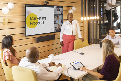 A photograph of a woman standing in a meeting space with a Google Meet Series One Medium Room Kit. She is standing in front of a large screen which displays an active Google Meet call and a Google Slides presentation. People are sitting at the table in front of her.