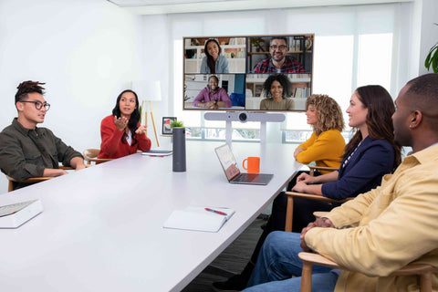 A photograph of a meeting room with several people sitting around the table and an active Google Meet call on the display at the front of the room. There is a Logitech Rally Bar mounted below the display. On the table there is a Logitech Sight and Logitech Tap along with a laptop, mug and two notepads.