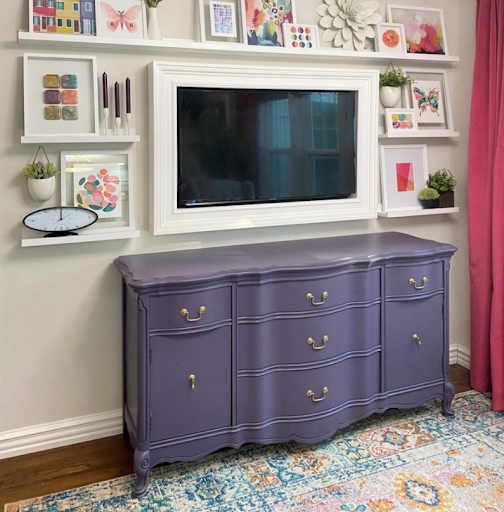 A quirky room decorated with brightly-colored wall art, a rug, and a purple-painted dresser