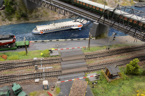  A model railroad set with a river, bridge, train tracks, a boat, grass, trees, and a railroad crossing