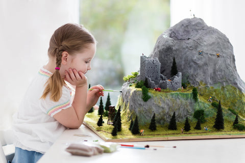 A little girl painting a mountain model on a table with trees and rock climbers, with a window in the background
