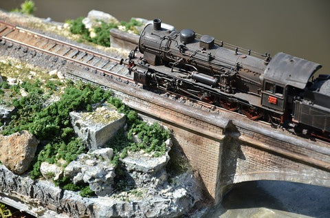 A diorama of a weathered model steam train crosses a bridge toward a rocky outcrop full of fake vegetation 