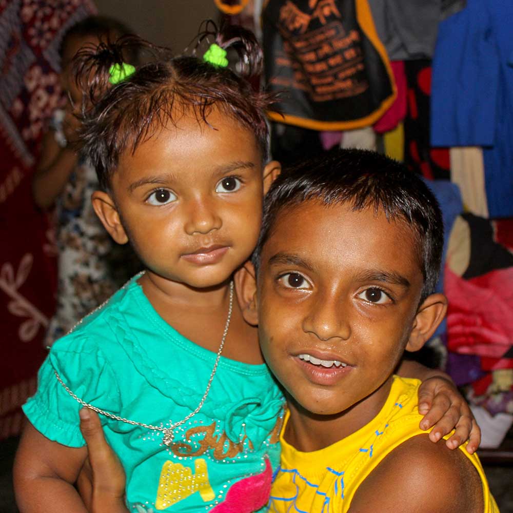 2 children from Bangladesh embrace one another, dark glowing skin, boy smiling, girl wearing cute ponytails