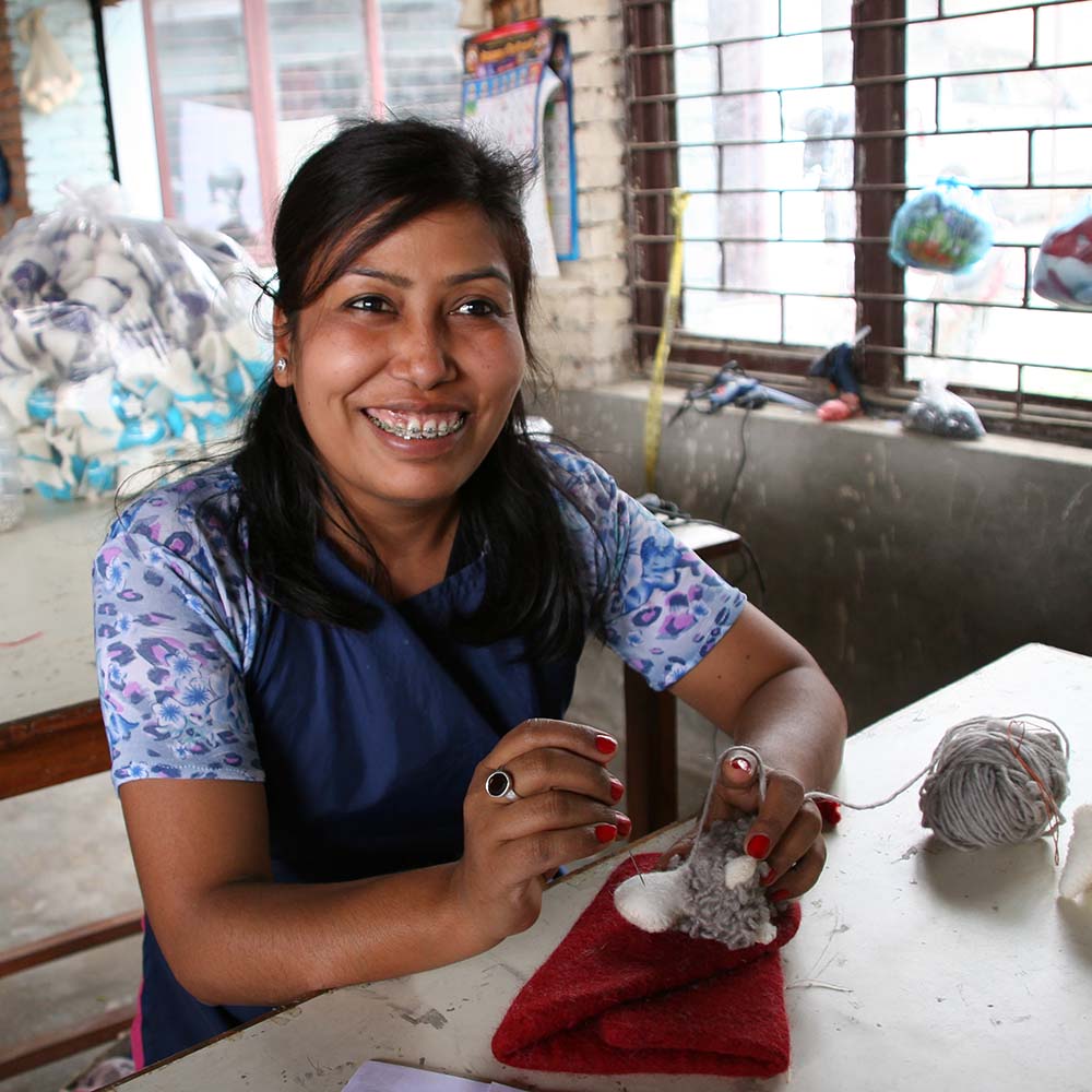 smiling Nepalese woman with braces on her teeth, sewing a fair trade toy for do good shop artisan partner
