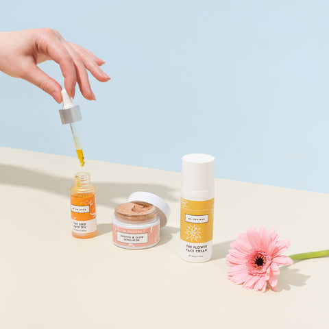 Three Be Fraiche products on beige and baby blue background. From left to right: a beautiful woman's hand holding The Seed Face Oil dropper with golden oil dripping from the tip, Smooth and Glow Exfoliator with lid open showing the lucious scrub texture, The Flower Face Cream and a pink flower.