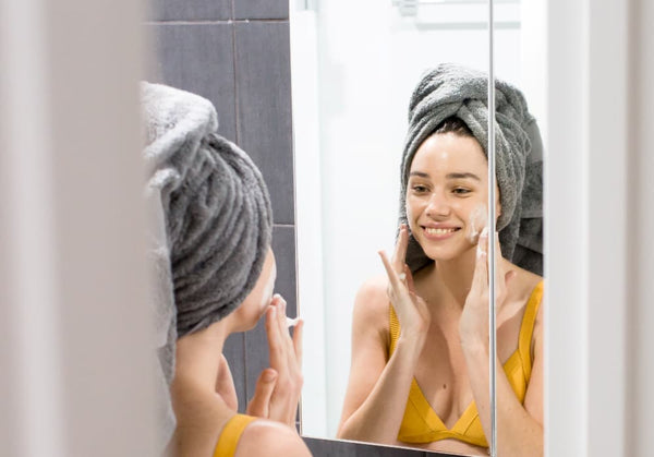 Image of a young woman using Be Fraiche Tea Cleanser on her face, smiling in front of a mirror