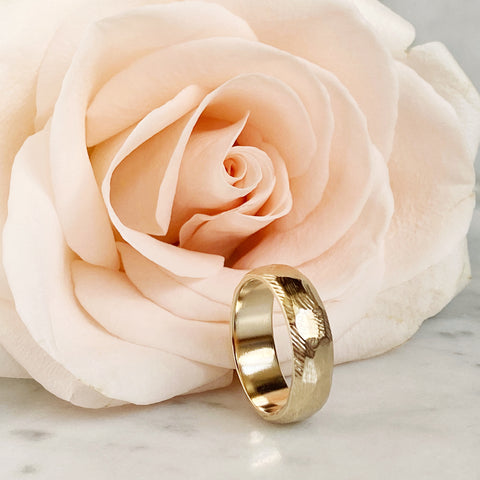 A faceted, textured gold wedding band standing on its edge, in front of a soft-peach colored rose