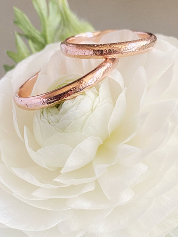 Two textured rose-gold thin wedding bands resting atop a white flower bloom