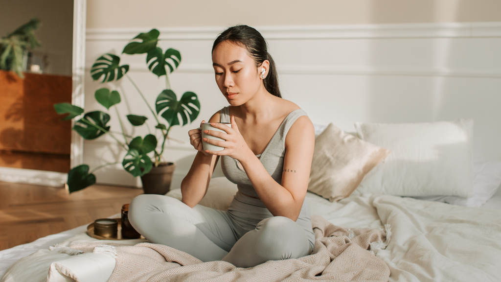 woman sits on a bed drinking from a mug in the morning light