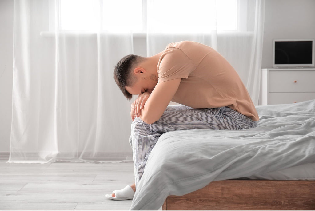 man sits on the edge of his bed with his head resting on his knees