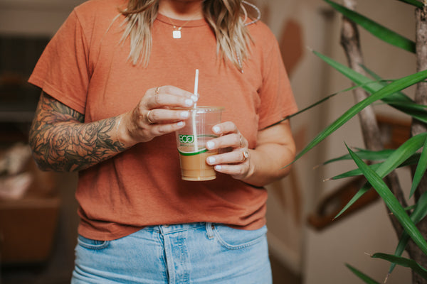 Jess wearing terracotta-colored shirt holding an iced coffee in front of plant. 