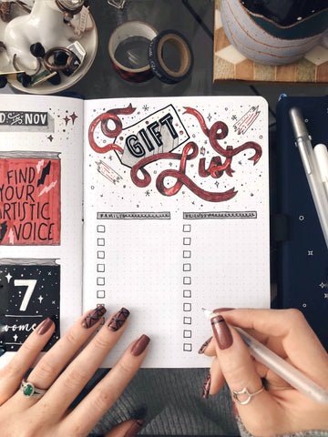An open bullet journal with the words "Gift List" at the top, with two female hands resting on the pages and holding a pen.