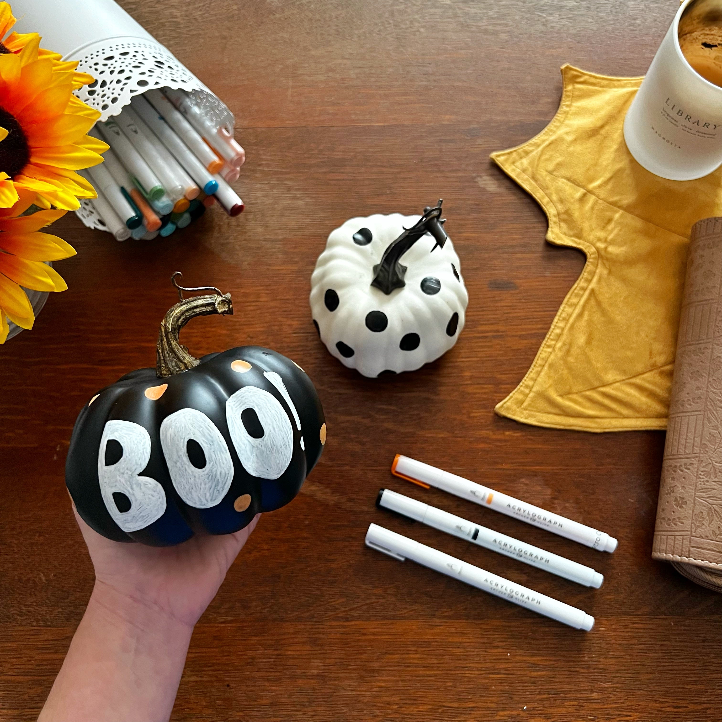 Picture of two artificial pumpkins embellished with Acrylograph pens. One with “Boo!”, and one with polka dots. Acrylographs scattered around.