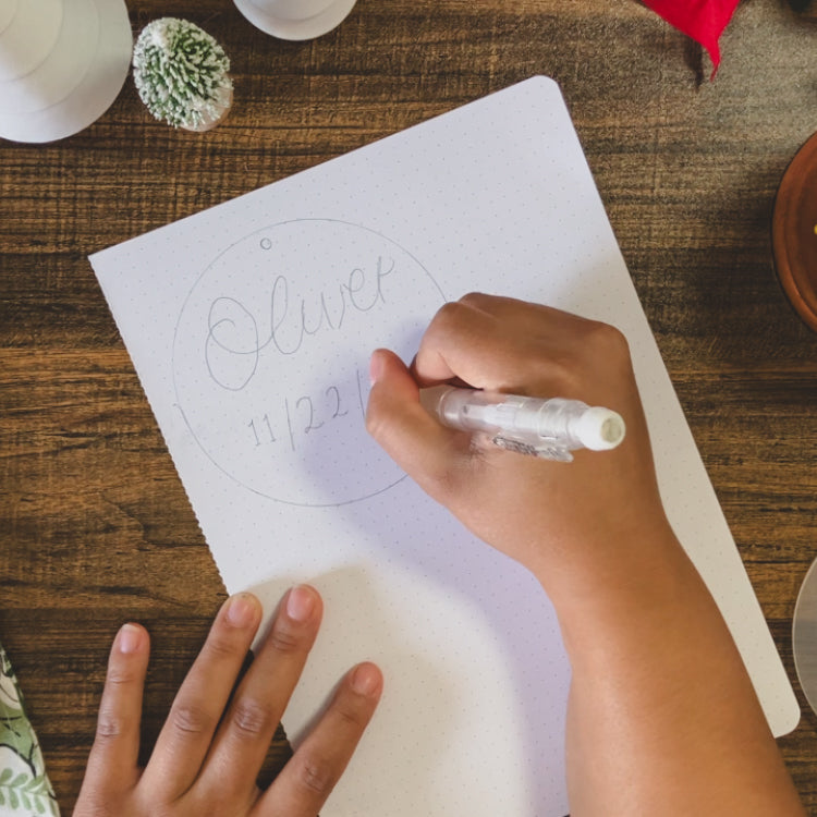 Emily is sketching an ornament design onto a piece of paper. The ornament is the name Oliver in calligraphy with a birth date below it.