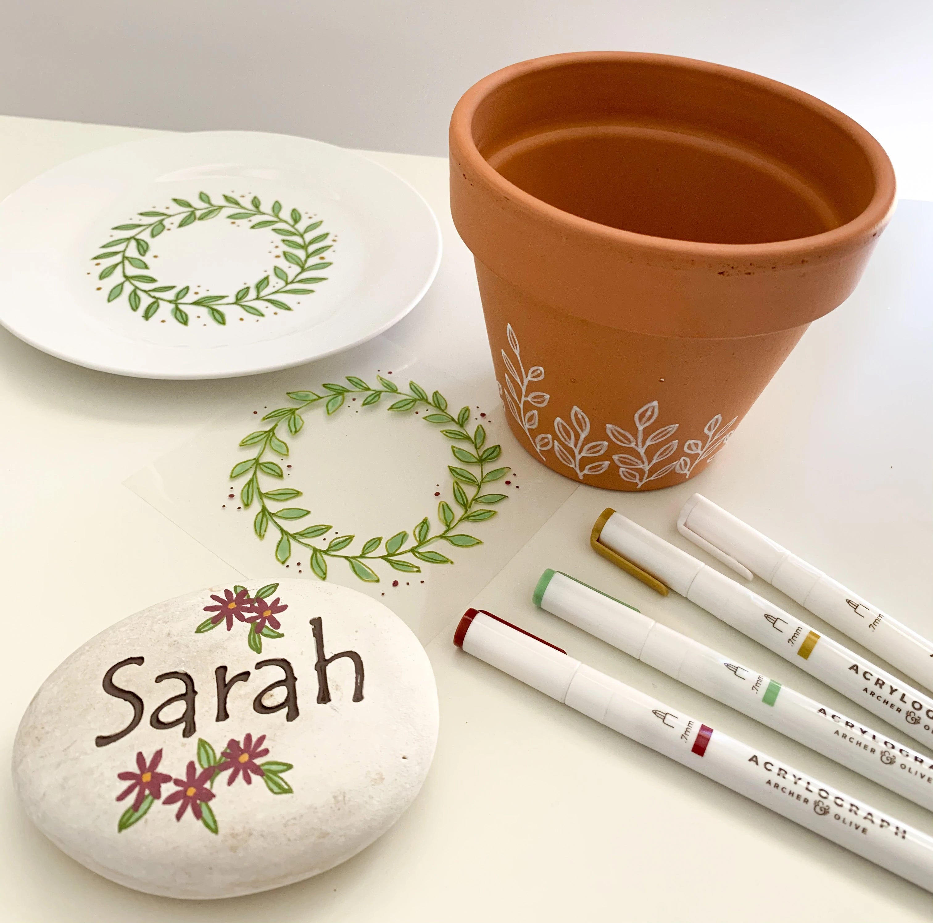 image of a planter, plate, and rock with  floral acrylograph designs all over, and the corresponding acrylograph pens on the side