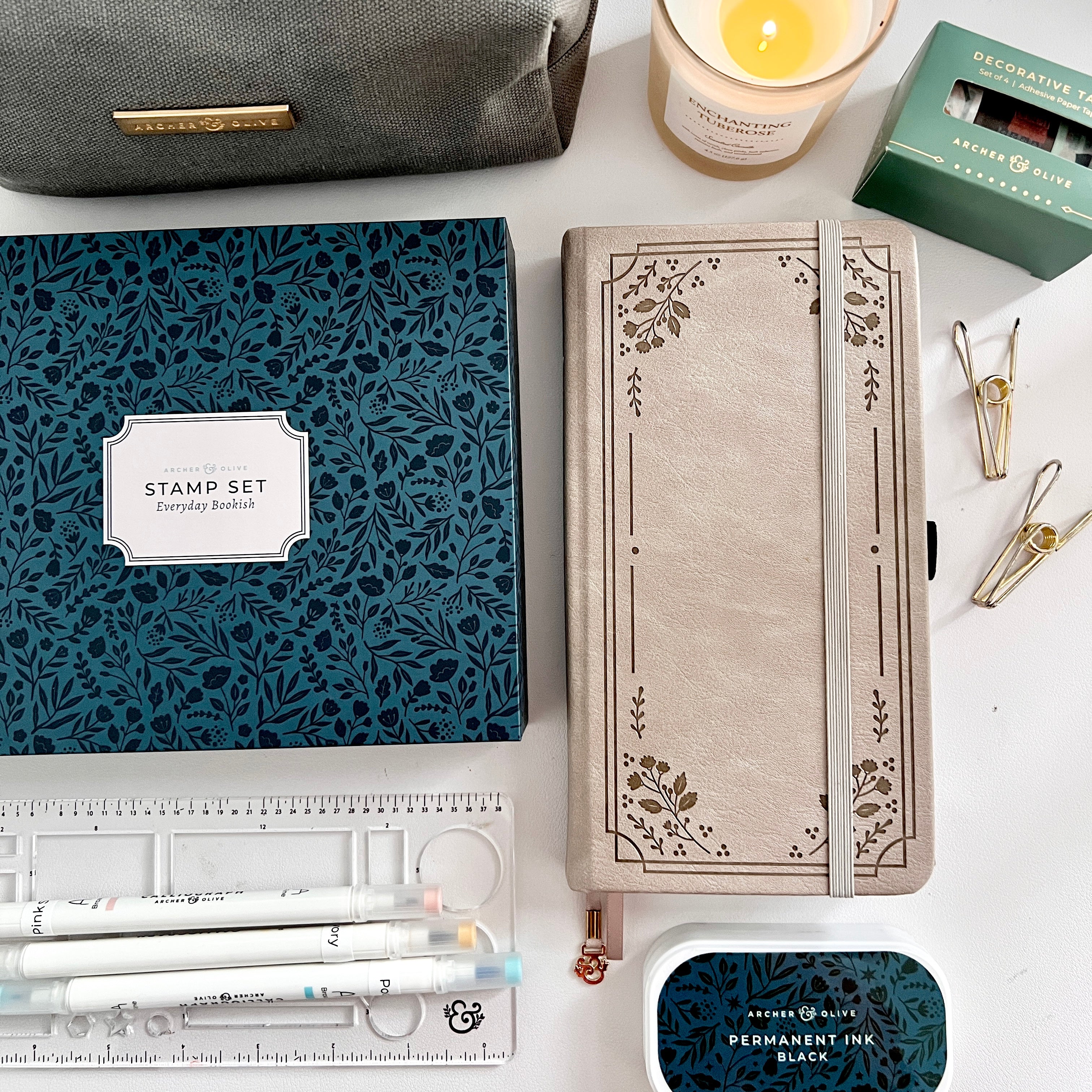 Flat lay image showing a travelers size bullet journal, stencil, stamp set, pens, a pencil pouch, a washi tape set, and lit candle