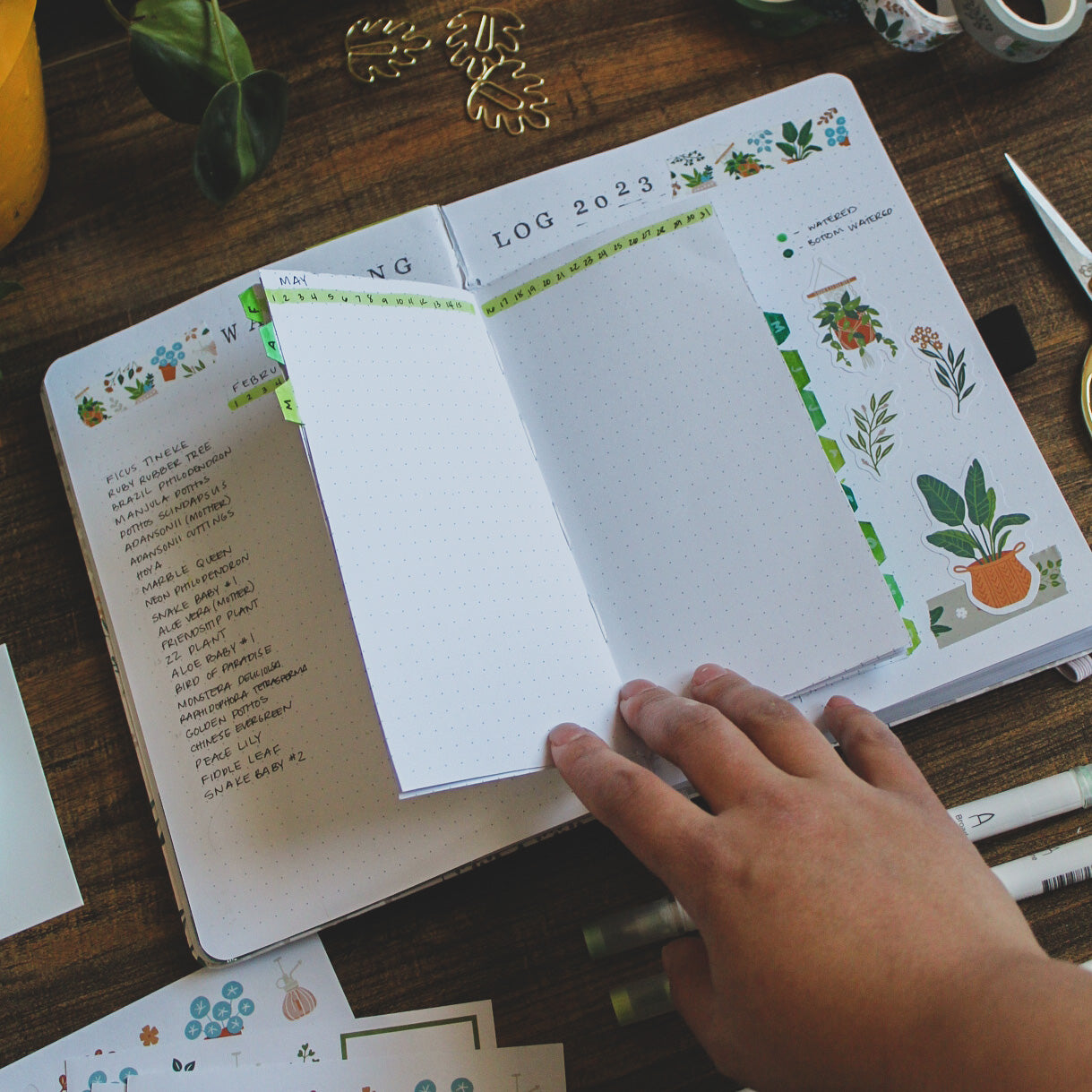 A dot grid journal lying open on a wood desk surrounded by journaling supplies. The pages are filled with a watering log spread for plant care made with dutch doors and other decorative elements.