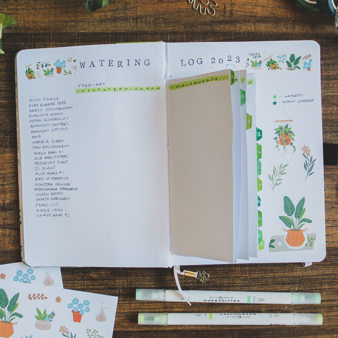 A journal is lying open on a dark desk surrounded by stationery supplies. The bullet journal spread shows a watering tracker for plant care using dutch doors.