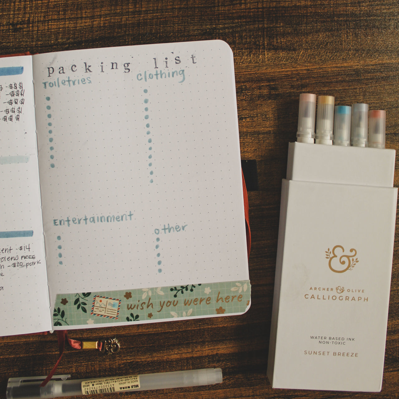 A dot grid notebook is lying open on a desk with one page visible. On the desk it is surrounded by other stationery elements like Calliograph brush pens. On the page is a packing list bullet journal spread.