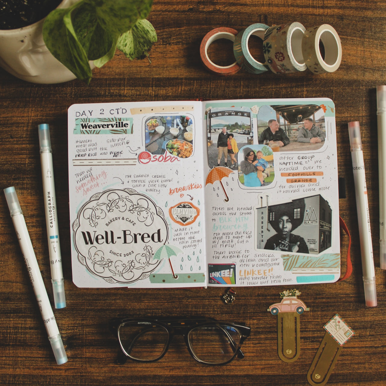 A dot grid notebook is lying open on a dark table. On the pages are travel memories spread including lettering, logos from businesses, stickers, and photos from the trip. On the table the notebook is surrounded by various stationery elements.