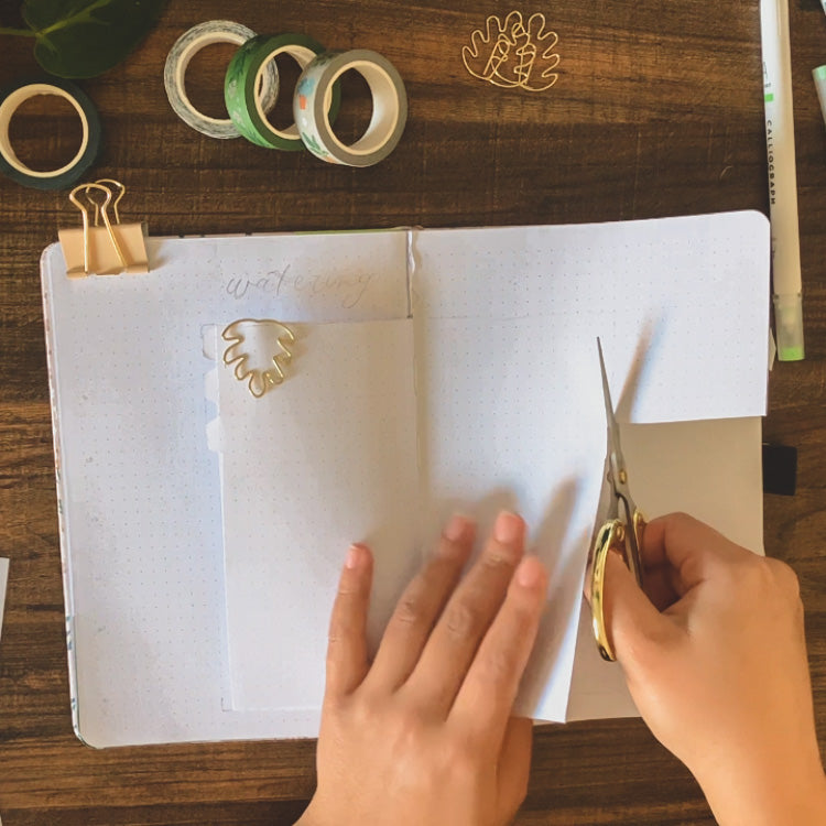 A dot grid notebook is lying open on a dark background surrounded by stationery supplies. Emily is cutting out a dutch door spread in the journal 