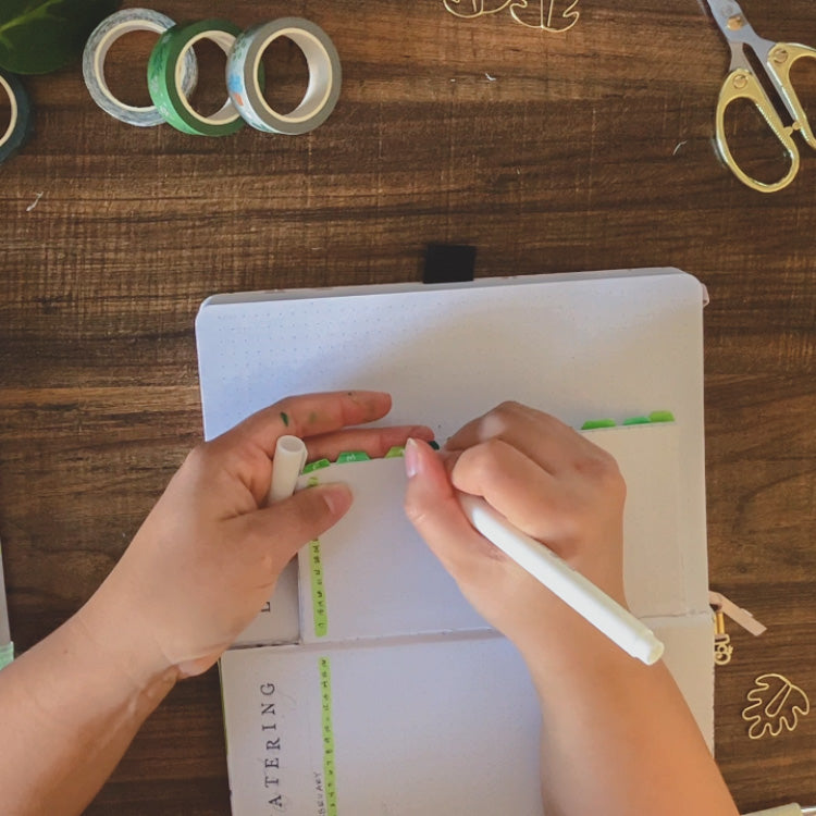 A dot grid notebook is open on a dark surface surrounded by journaling supplies. Emily is labeling the index tabs in the dutch door spread with a white acylograph paint pen.