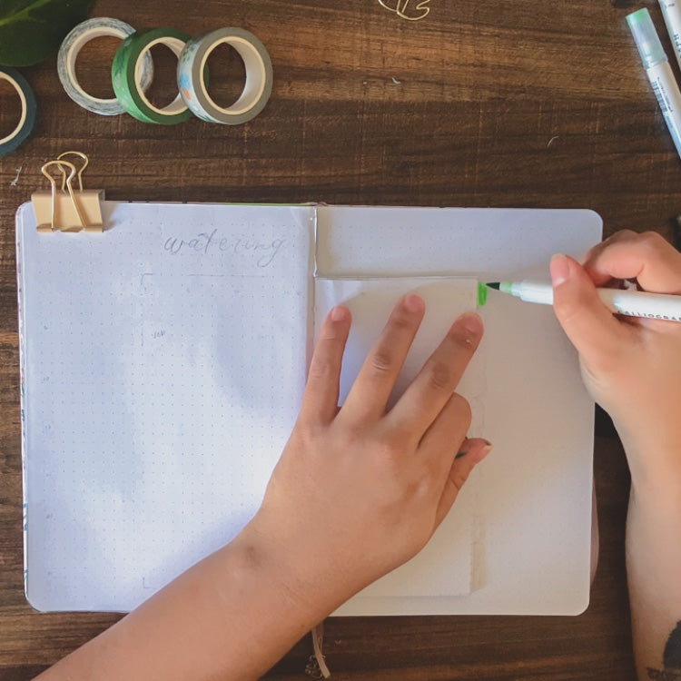 A dot grid journal is lying open on a dark surface. Emily is coloring in the index tabs on the dutch doors with green calliograph brush pens.