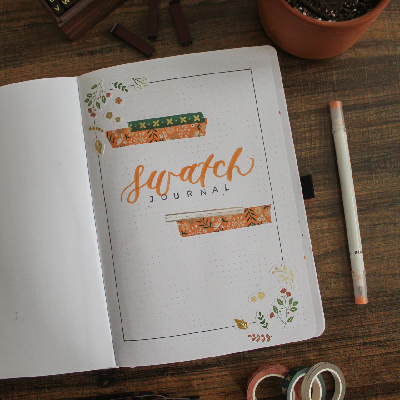 A notebook is lying open on a dark surface. On the page is the title "Swatch Journal" with some decorative stickers and washi tape. On the desk around the notebook are stationery items.