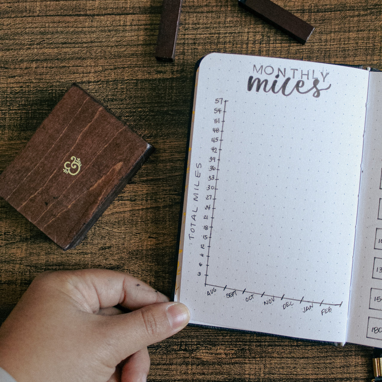 A hand is holding open a pocket sized notebook. On the notebook is a line graph set up to track how many miles are ran each month.