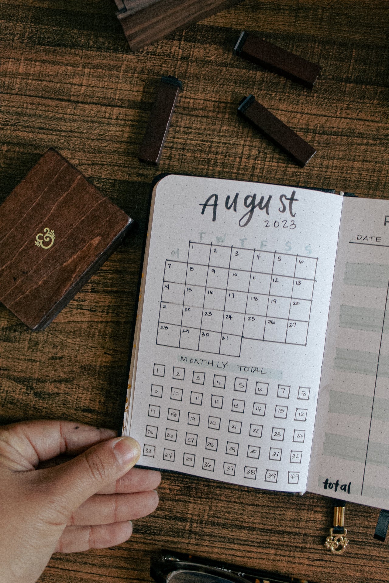 A pocket notebook is lying open on a desk. On the page is a small calendar spread for August 23 and a running goal tracker for the month.