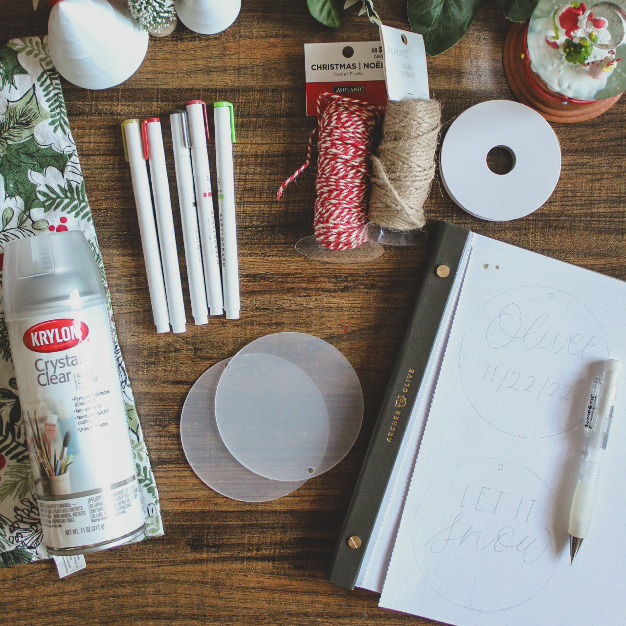 The supplies for these DIY Christmas Ornaments are laid out on a desk with decorative Christmas elements around them. The supplies are some blank acrylic rounds, Archer and Olive Acrylograph Paint Pens, twine or ribbon, a pencil, sketch paper, and some sealant spray.