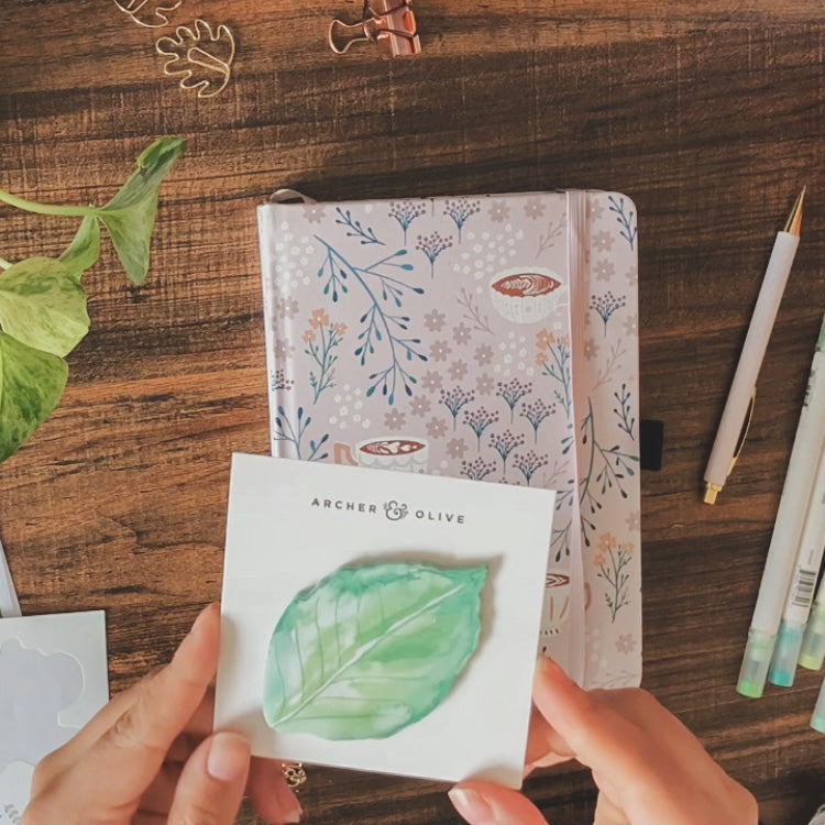 A notebook is lying on a desk surrounded by stationery elements. Emily is holding up a leaf-shaped sticky note pad found in the Growing Seasons monthly kit.