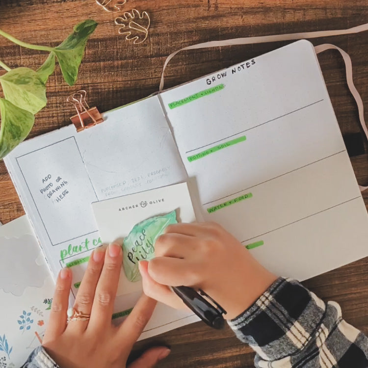 A white dot grid notebook is open on a desk surrounded by plant themed stationery items. Emily is writing the name of a plant, peace lily, onto a sticky note that looks like a plant leaf.