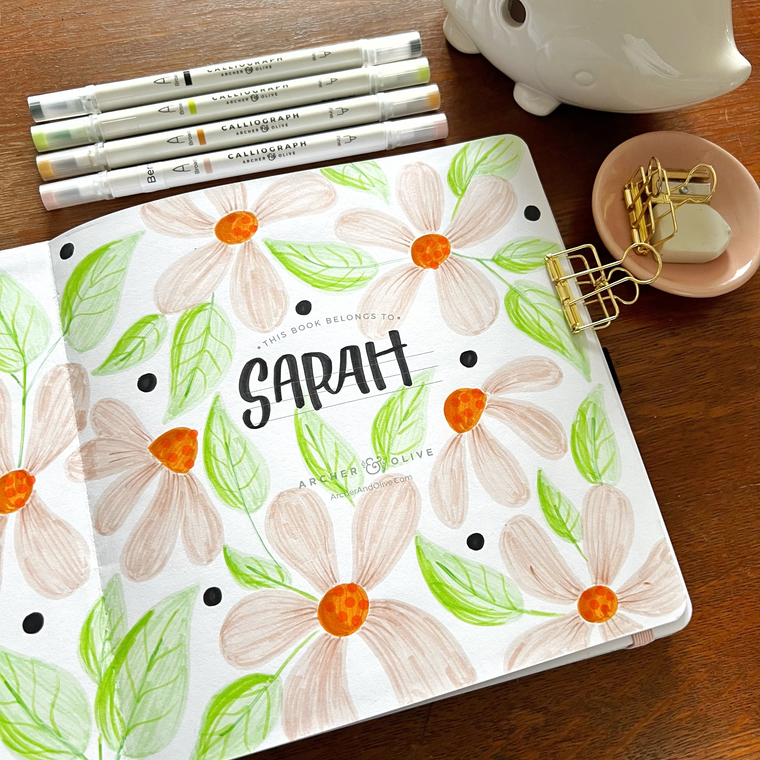 Open notebook with floral doodles covering page. Calliograph pens and various stationery surrounding