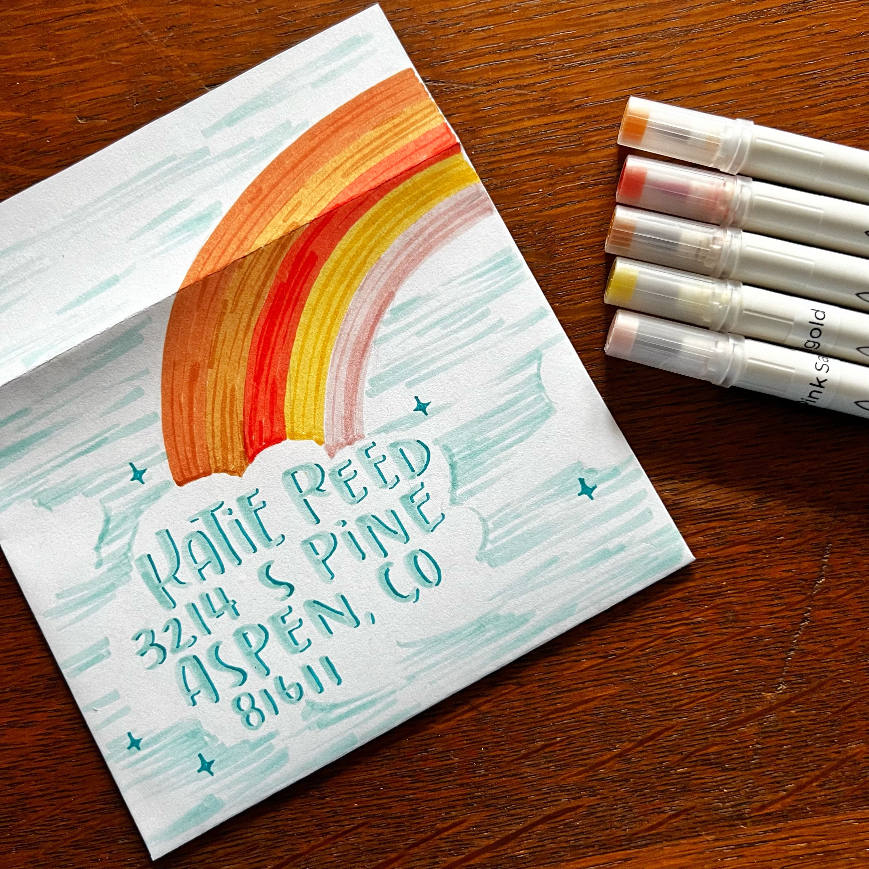 Picture of envelope decorated with rainbow and cloud. Calliograph pens next to envelope.